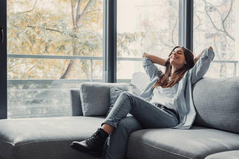 Woman relaxing comfortably on a couch in a cozy, well-humidified home, enjoying balanced indoor air quality near large windows.