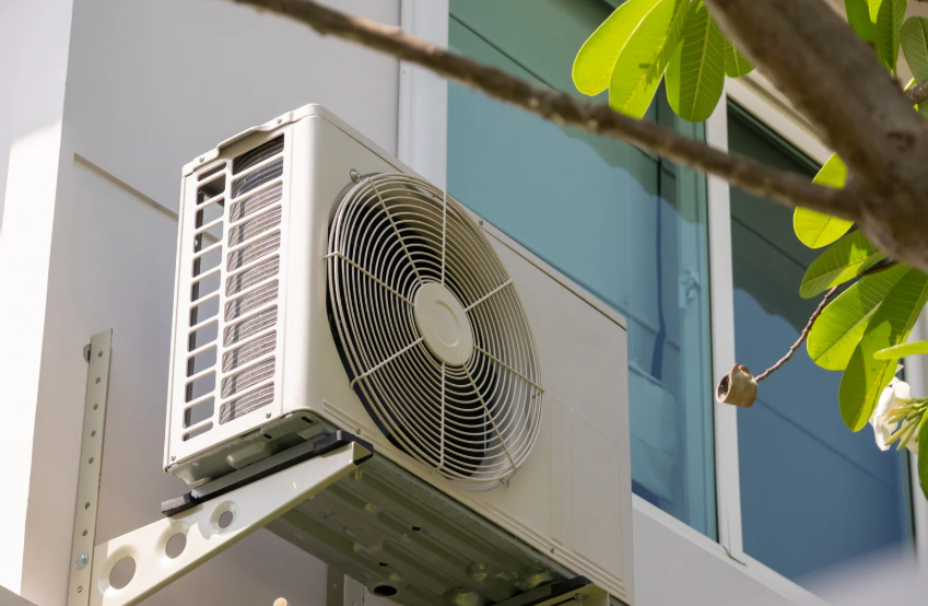 An outdoor air conditioning unit mounted on a bracket outside a building near a window, with leaves and a tree branch in the foreground.