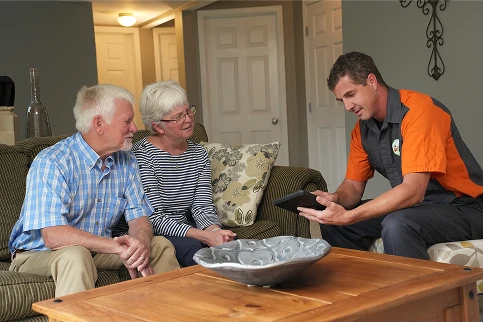 An elderly couple sits on a couch while a worker in a uniform shows them information on a tablet in a living room.