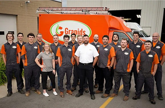 A group of employees wearing uniforms stands in front of an orange work van and building with a "Granpids" logo.