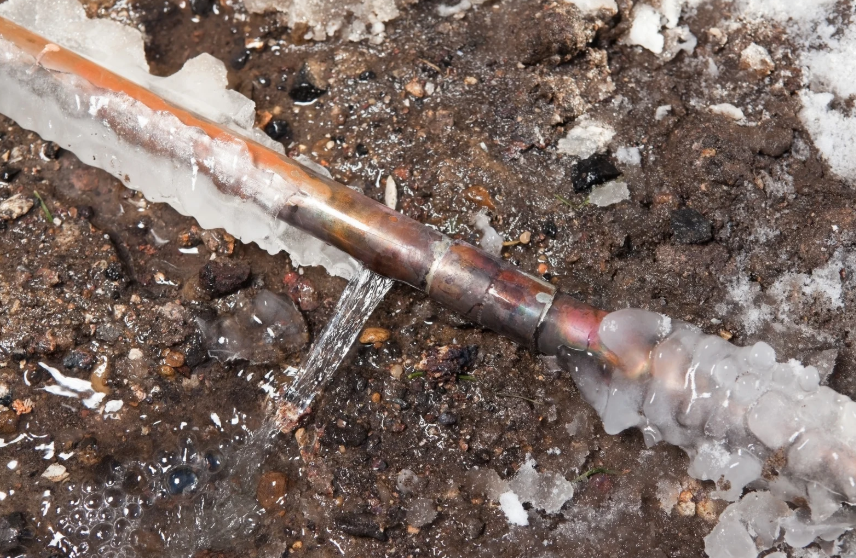 A copper pipe with a crack leaks water onto muddy ground, surrounded by ice and snow.