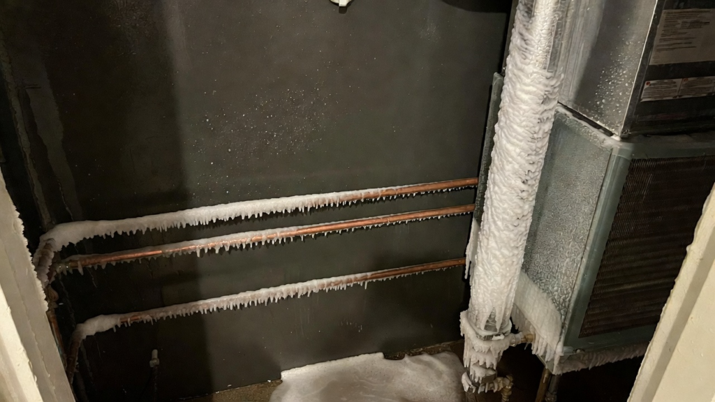 Frozen pipes and HVAC system components covered in thick ice and frost, with ice buildup on the floor and walls in a utility room.