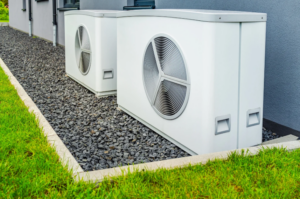 %Focuskw% | Grapids Home Services Two white outdoor heat pump units are installed on a bed of gray gravel next to a building, bordered by a strip of green grass.