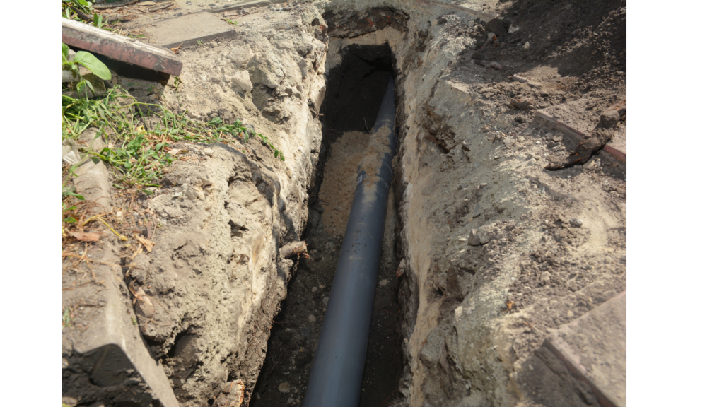 A grey plastic pipe is installed in a narrow trench dug into the ground, with soil and grass visible along the edges.