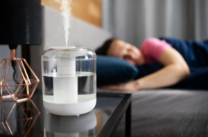 %Focuskw% | Grapids Home Services A cool mist humidifier emitting vapor sits on a bedside table while a person sleeps in the background.