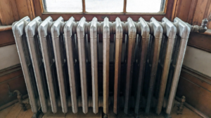 %Focuskw% | Grapids Home Services A cast iron radiator with visible discoloration and aging, positioned in front of a wooden-framed window and wall.