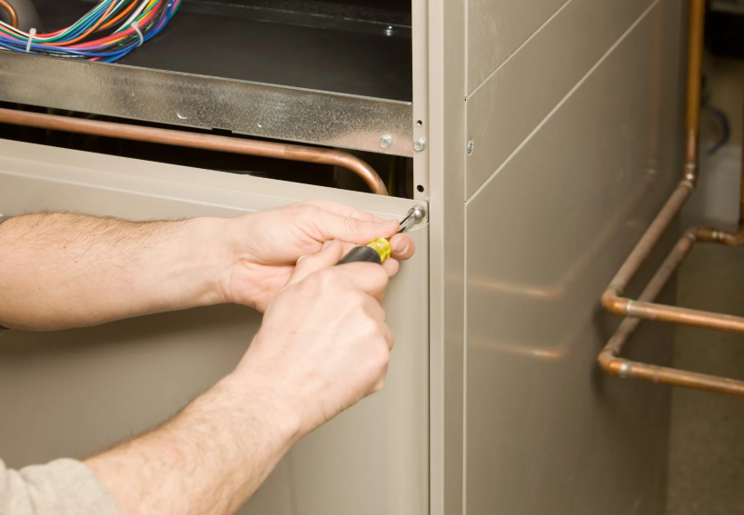 A person uses a screwdriver to remove or tighten a screw on the side panel of a metal appliance, with wires and copper pipes visible inside.