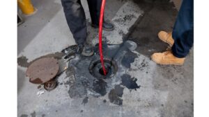 %Focuskw% | Grapids Home Services Two people stand near an open manhole cover while feeding a red hose or cable into the hole on a dirty concrete surface.