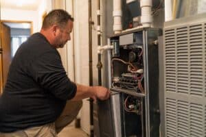 %Focuskw% | Grapids Home Services A man wearing a black shirt repairs or inspects an open furnace unit in a residential utility room.