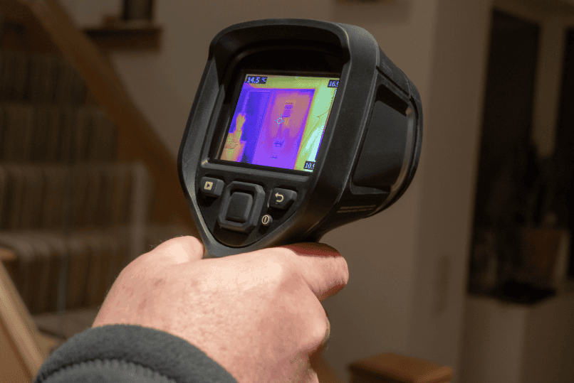 A person holds a thermal imaging camera, displaying a heat map of a staircase and wall area inside a house.