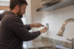 %Focuskw% | Grapids Home Services A man uses a screwdriver to install or repair an electrical outlet above a kitchen counter near a sink and faucet, mindful of safety practices often discussed when considering questions like how long does an HVAC system last.
