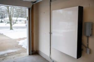 %Focuskw% | Grapids Home Services A large white battery storage unit is mounted on the wall of a garage, with snow visible outside through the open garage door—a reminder of how important efficient backup power can be, especially when considering how long an HVAC system lasts in challenging climates.